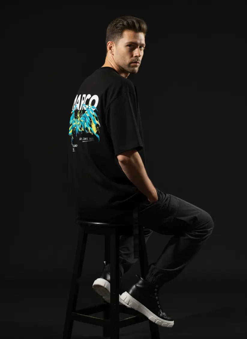 Man wearing a black t-shirt with a colorful graphic design sitting on a stool against a dark background